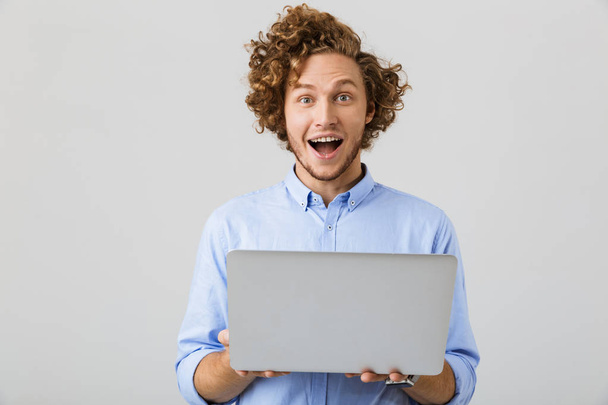 Portrait of an excited young man wearing shirt standing isolated over gray background, holding laptop computer - Fotó, kép