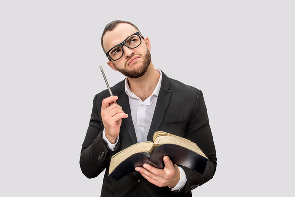 Thoughtful and serious young man holds book and pen. He looks up though glasses. Young man wears suit. Isolated on white background. - Photo, image