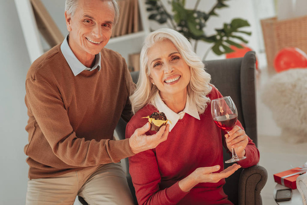 Smiling senior spouses siting in living room with wine and cake - Fotó, kép