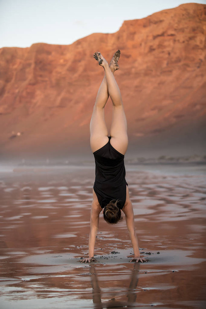 Вид на здоровую женщину, показывающую стойку на руках, занимающуюся йогой на пляже, стоящую на мокром песке
 - Фото, изображение