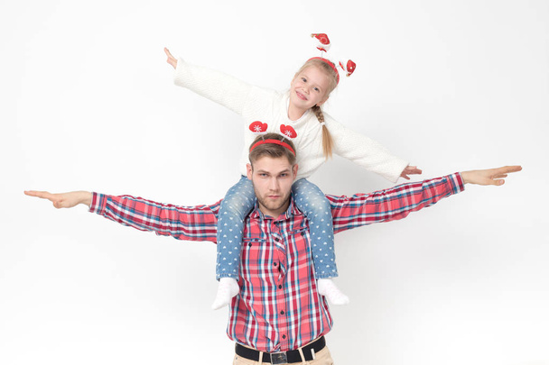A little girl is sitting at her fathers back. Family in funny Christmas headbands on a white background. - Фото, изображение