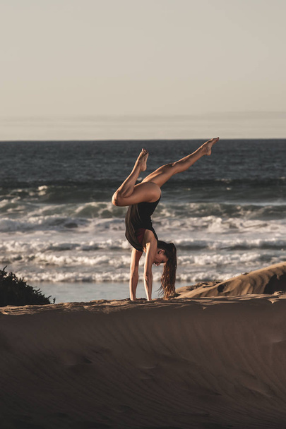 Side view of strong woman in yoga handstand on sandy shore with waves of ocean on background - Foto, imagen