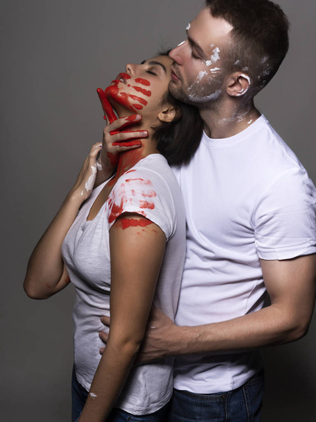 Beautiful couple of artists wearing jeans and white t-shirts soiled with white and red paint sensually hug each other on gray. Palm prints painted on their faces. Concept, fashion design. Copy space - Foto, Bild