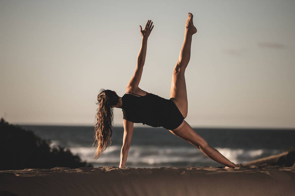 Back view of slim woman in black bodysuit doing side asana training yoga alone on sandy beach - Фото, изображение