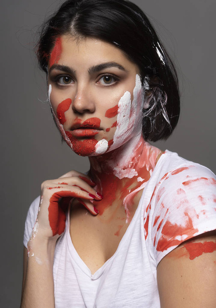 Beautiful brunette girl wearing a white t-shirt stained with paint touches her face and neck with her hand. Handprint of the palm and strokes of paint on her face. Conceptual, fashion design - Foto, imagen
