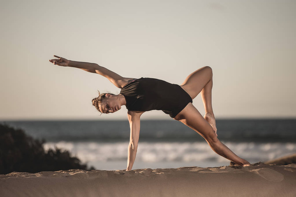 Beautiful slim woman in black bodysuit training yoga pose on sandy beach with ocean landscape - Φωτογραφία, εικόνα
