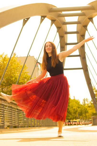 Молодая стройная красивая женщина с длинными каштановыми волосами танцует и улыбается на открытом воздухе в черной рубашке и терракотовой (красная груша) длинной юбке. Спорт и здоровая концепция
. - Фото, изображение