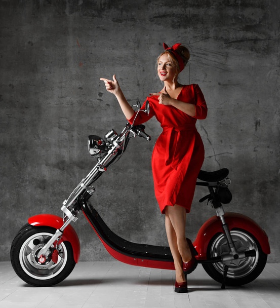 Woman ride sit on motorcycle bicycle scooter pinup retro style pointing finger laughing smiling red dress  - Фото, изображение