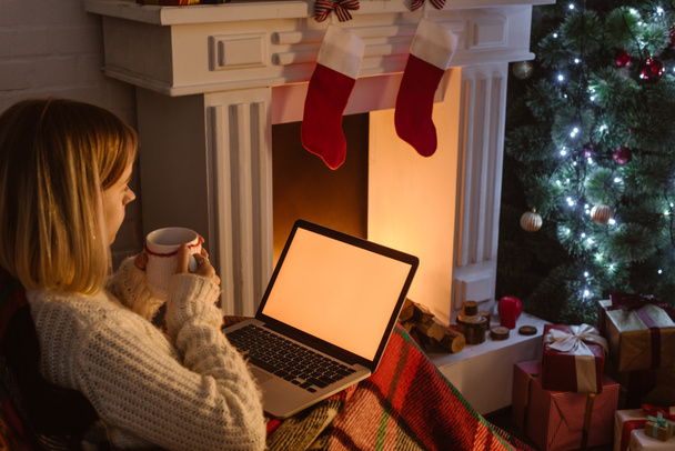 young woman holding cup of cocoa and using laptop with blank screen at christmas time - Photo, Image