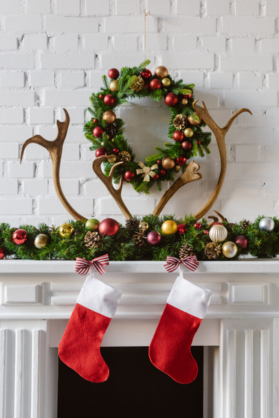 εορταστικό Χριστουγεννιάτικο στεφάνι με διακοσμήσεις, κάλτσες και ελάφια κέρατα πάνω από την κορνίζα τζακιού εστιών - Φωτογραφία, εικόνα