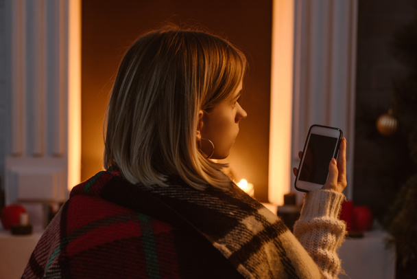 молода жінка в картатому ковдрі використовує smaprtphone з порожнім екраном вдома
 - Фото, зображення