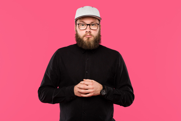 serious bearded male hipster in cap hat and eyeglasses looking at camera isolated on pink  - 写真・画像