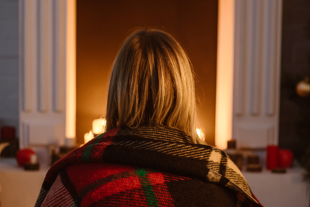 back view of woman in checkered blanket sitting near fireplace at home - Photo, Image
