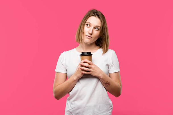 dreamy girl holding disposable cup of coffee isolated on pink - Zdjęcie, obraz