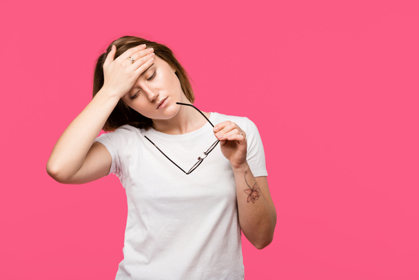 εξαντληθεί νεαρή γυναίκα κρατώντας γυαλιά ενώ έχοντας πονοκέφαλος που απομονώνονται σε ροζ - Φωτογραφία, εικόνα