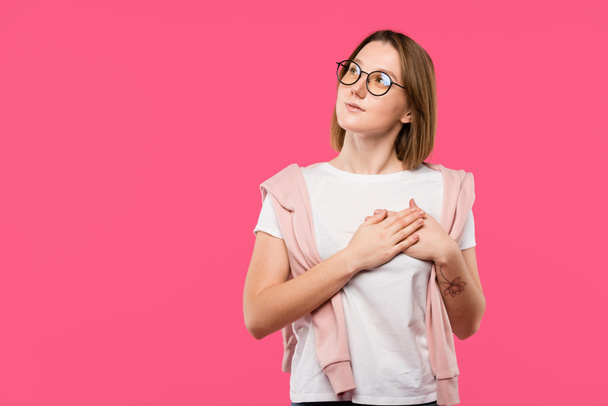 dreamy girl in eyeglasses holding hands on chest and looking away isolated on pink - Zdjęcie, obraz