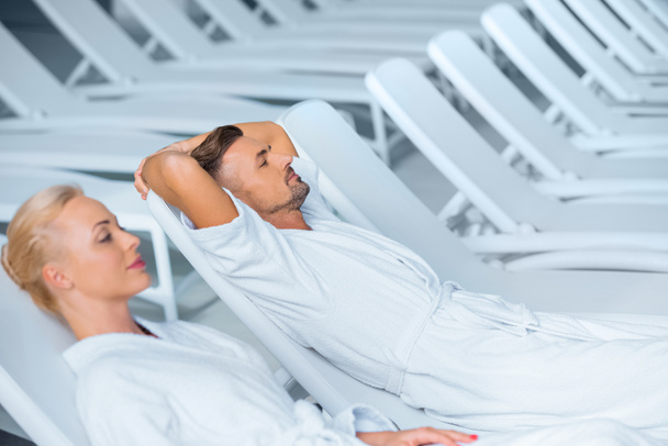 Couple relaxing on deck chairs in white bathrobes  - Photo, Image