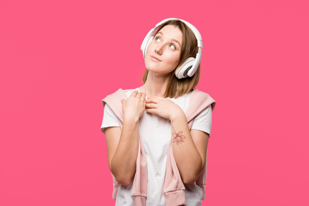 joven mujer de ensueño en auriculares escuchando música aislada en rosa
 - Foto, Imagen