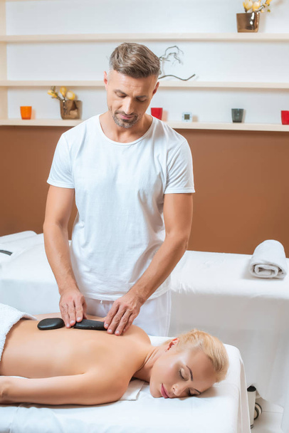 Handsome therapist putting hot stones on back of woman  - Photo, Image