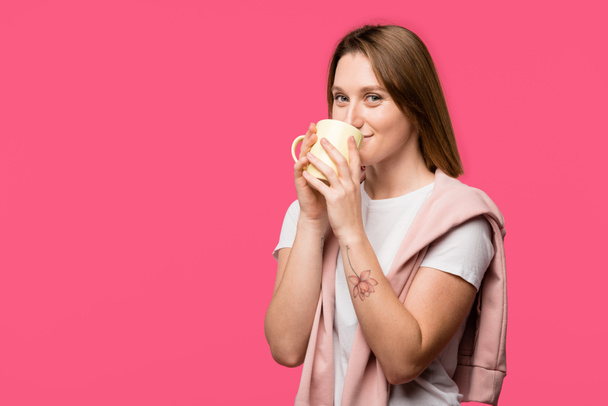 молода жінка п'є з чашки і дивиться на камеру ізольовано на рожевому
 - Фото, зображення