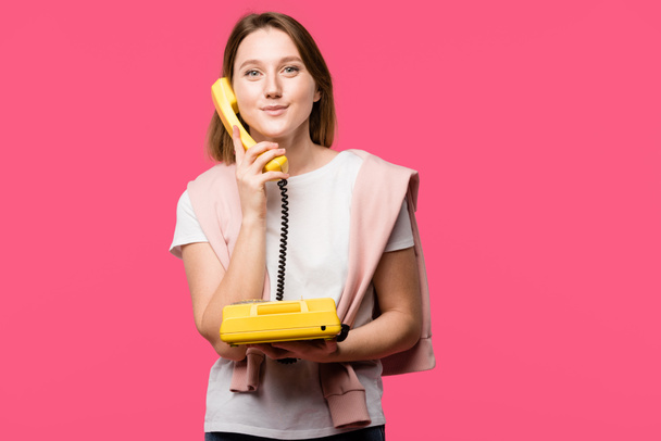 beautiful young woman talking by rotary phone and smiling at camera isolated on pink - Fotó, kép