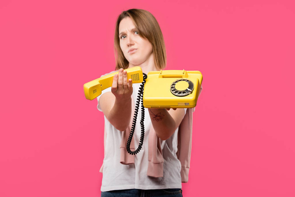 mujer joven con la cara frunciendo el ceño sosteniendo teléfono rotatorio amarillo aislado en rosa
 - Foto, Imagen