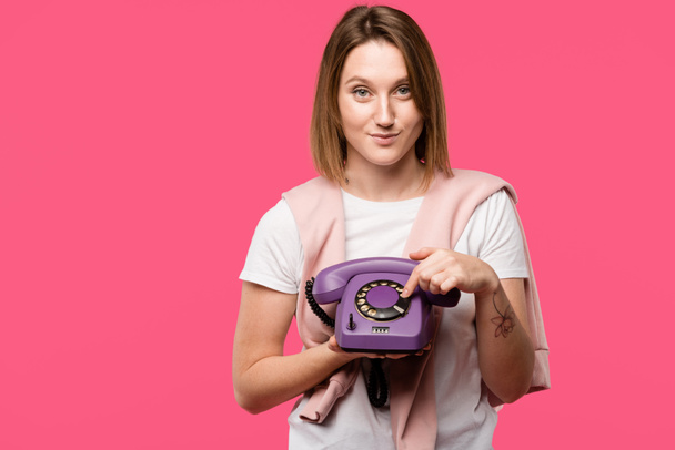 hermosa mujer joven sosteniendo el teléfono giratorio y sonriendo a la cámara aislada en rosa
 - Foto, Imagen
