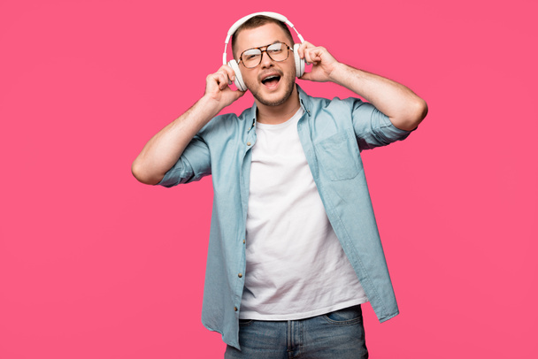 felice giovane uomo con gli occhiali che ascolta musica in cuffia e sorride alla fotocamera isolata su rosa
  - Foto, immagini