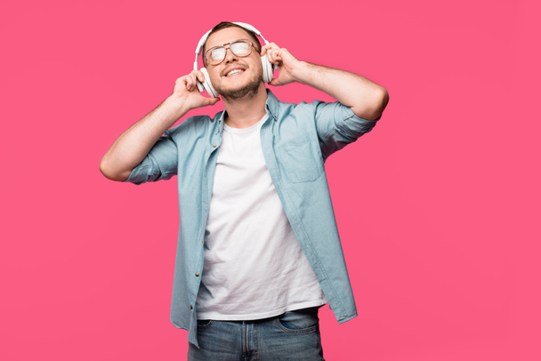 χαρούμενος νεαρός άνδρας σε γυαλιά ακούγοντας μουσική στα ακουστικά που απομονώνονται σε ροζ  - Φωτογραφία, εικόνα