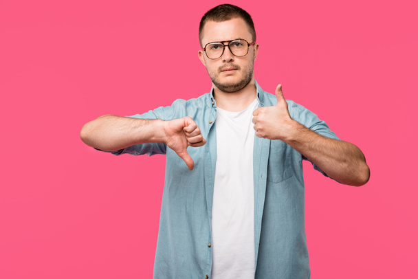 nejistý muž v brýlích ukazuje palec nahoru a palec dolů izolované na růžové - Fotografie, Obrázek