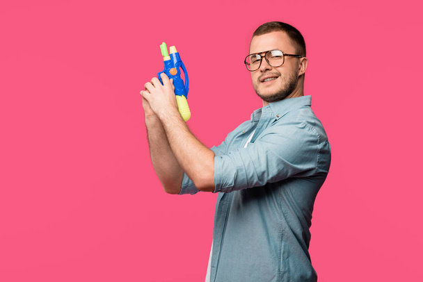 νεαρός άνδρας σε γυαλιά κρατώντας όπλο παιχνίδι και να χαμογελά στη φωτογραφική μηχανή που απομονώνονται σε ροζ  - Φωτογραφία, εικόνα
