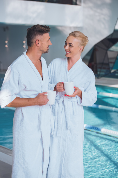 smiling couple in white bathrobes standing near swimming pool and holding cups - Foto, immagini
