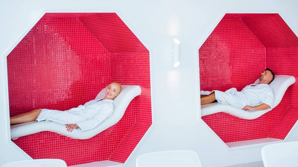 blonde vrouw en knappe man in de witte badjassen rustend op de ligstoelen in mozaïek achthoeken - Foto, afbeelding