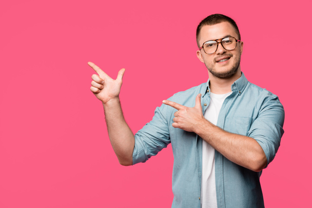 happy young man pointing with fingers at copy space and smiling at camera isolated on pink - Fotografie, Obrázek