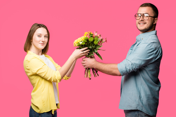 boční pohled na šťastný mladý pár drží kytici a usmívá se na fotoaparát izolované na růžové - Fotografie, Obrázek