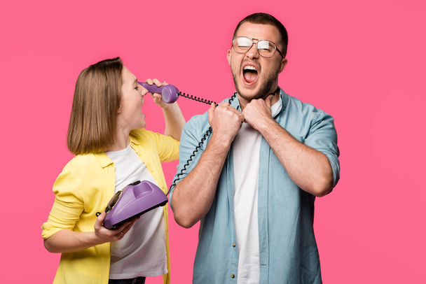 young woman talking by rotary phone and looking at man with wire around neck screaming isolated on pink - Foto, imagen
