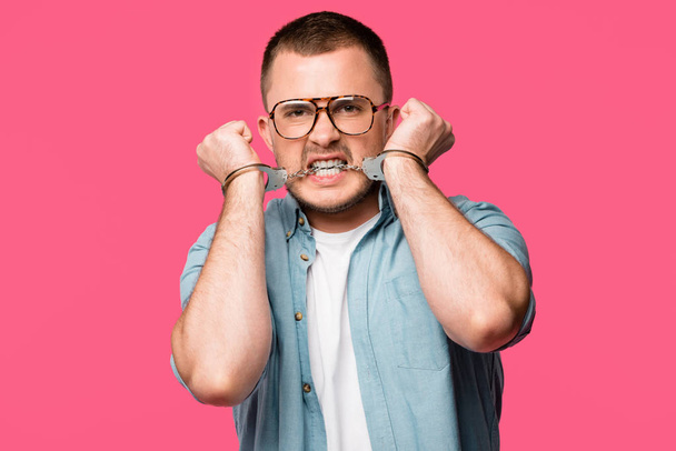 aggressive young man in eyeglasses biting handcuffs and looking at camera isolated on pink - Photo, image