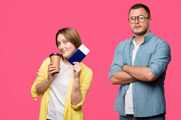 man in eyeglasses standing with crossed arms and looking at camera while smiling woman holding passport, boarding pass and coffee to go isolated on pink - Фото, зображення
