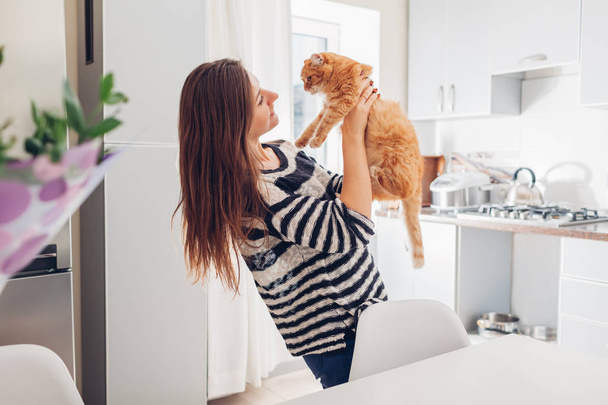 Young woman playing with cat in kitchen at home. Girl holding and raising red cat. Happy master having fun with her pet - Фото, изображение