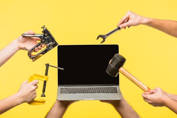 cropped shot of hands holding tools and laptop with blank screen isolated on yellow  - Photo, Image