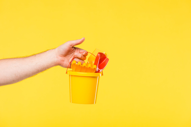 cropped shot of person holding plastic bucket with toys isolated on yellow - Zdjęcie, obraz