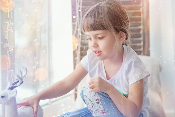 Gyermek játszó karácsonyi dekoráció. Kislány ül, az ablak mellett. Téli boldog ünnep fogalma. - Fotó, kép