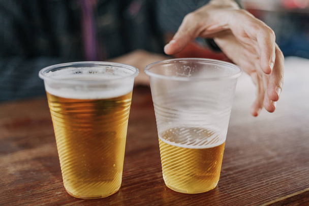 Twee beneveld eenmalig gebruik plastic glazen met bier staan naast elkaar op een donkere achtergrond. De man-hand reikt naar de incomplet - Foto, afbeelding