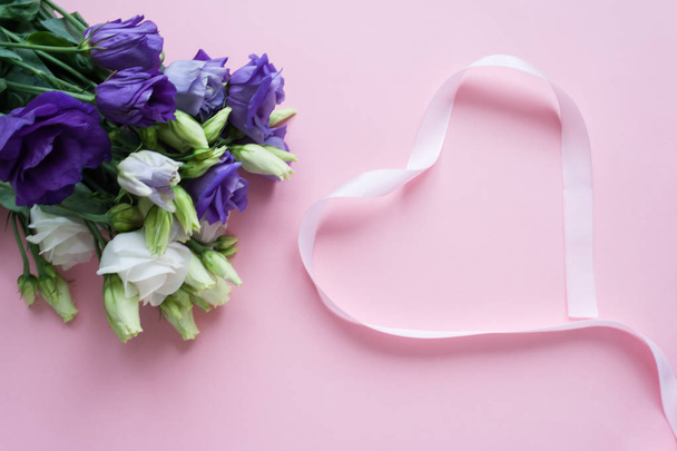 Frame of white Lisianthus and pink ribbon in the shape heart on pink background. Flat lay, top view - Fotografie, Obrázek
