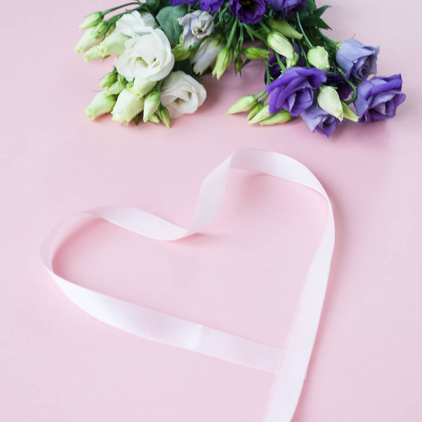 Frame of white Lisianthus and pink ribbon in the shape heart on pink background. Flat lay, top view - Photo, Image