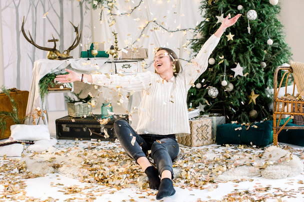 Pretty young woman in jeans and knitted white sweater sits near the Christmas tree and throws up golden confetti. Concept of Merry Christmas, New Year, family, holidays. - Fotografie, Obrázek