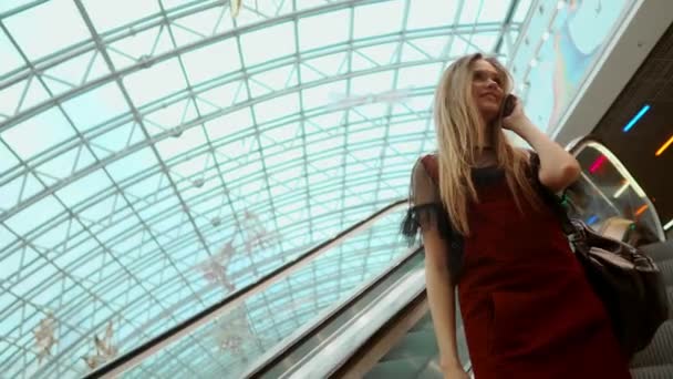 A beautiful girl descends the escalator and talks on the phone in the mall. - Séquence, vidéo