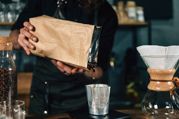 Detailní záběr z mladých mužů barista vylévá kávová zrna do sklenice a měření na digitální stupnici v kavárně. Barista s Tetovaný zbraně tmavé uniformě.    - Fotografie, Obrázek