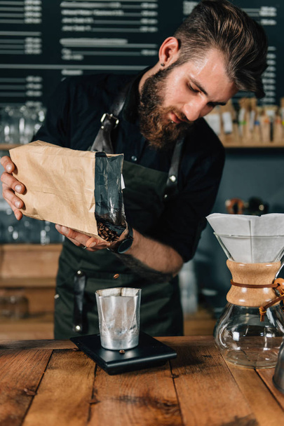 Close up van een jonge mannelijke barista koffiebonen in een glas gieten en meten op digitale weegschaal in coffeeshop. Barista met getatoeëerde armen dragen van donkere uniform.    - Foto, afbeelding