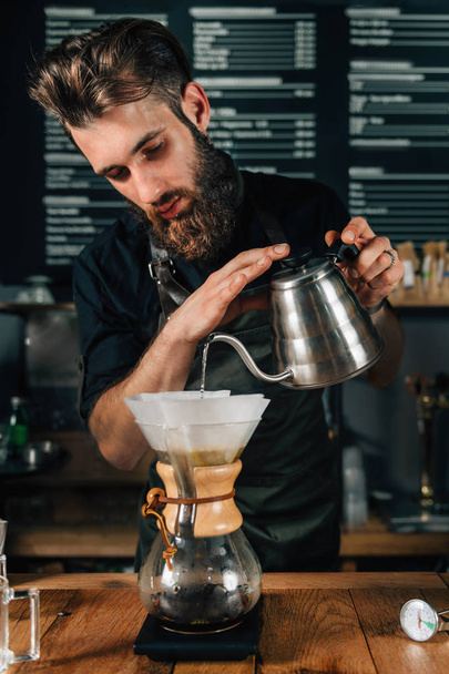 Mladý muž barista nalití vroucí vodu z konvice na kávu filtru. Barista s tmavé uniformě.     - Fotografie, Obrázek